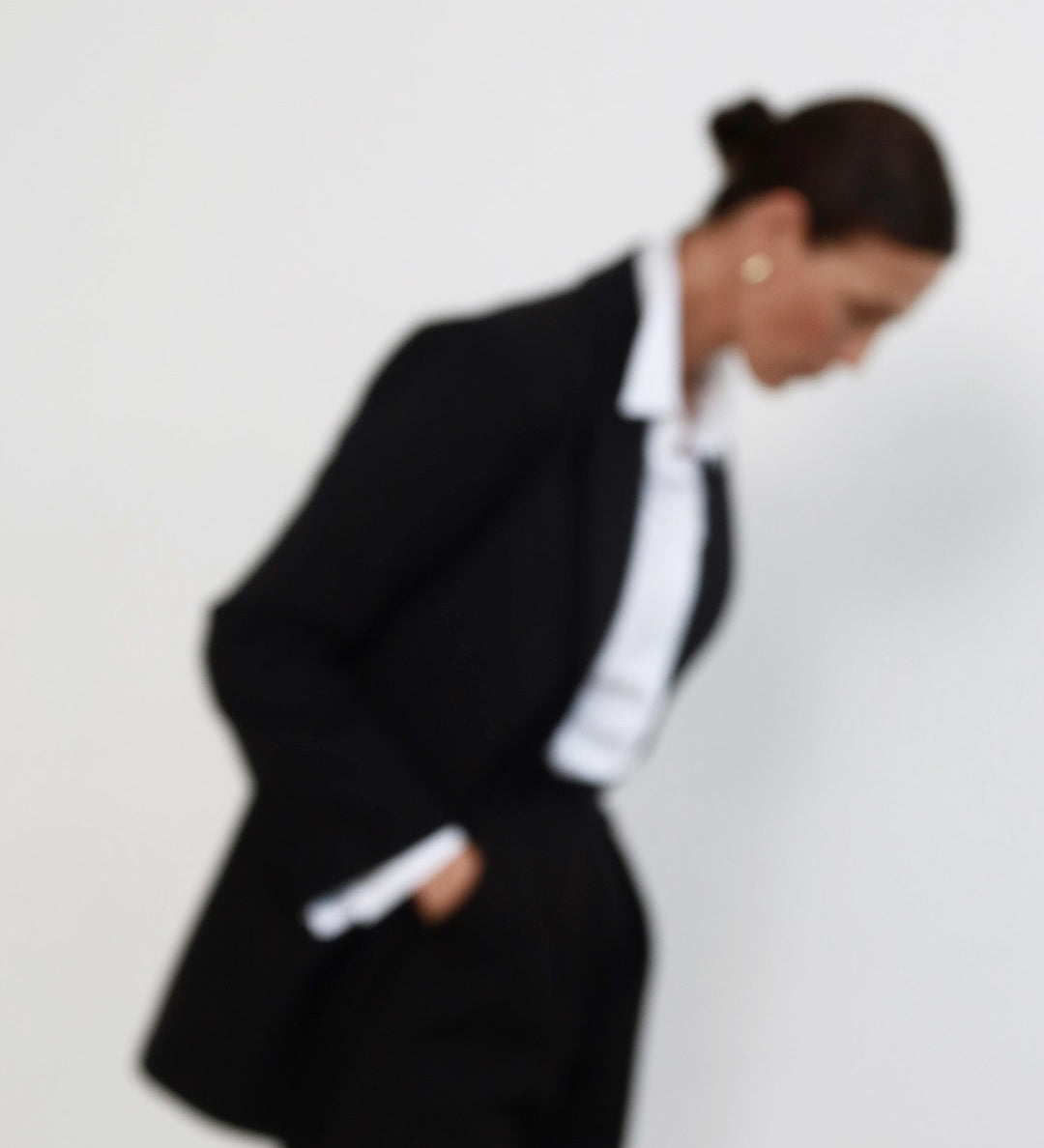 Out of focus image of a middle aged woman with pulled back dark hair wearing minimalist black suit with tailored blazer, and high waisted peg leg trousers