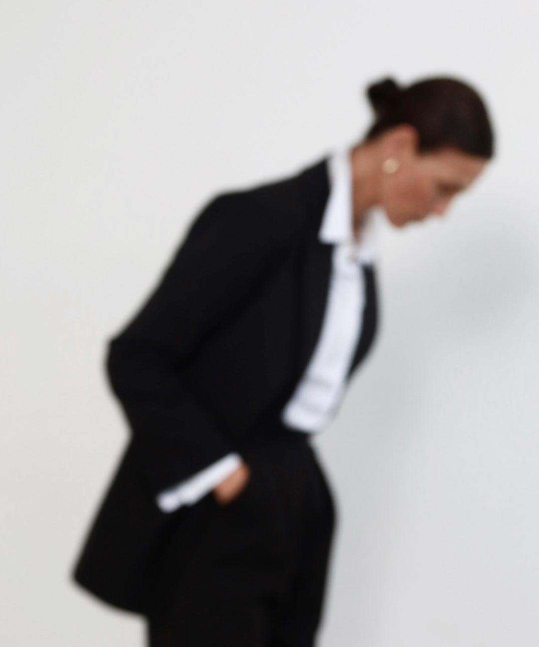 Out of focus image of a middle aged woman with pulled back dark hair wearing minimalist black suit with tailored blazer, and high waisted peg leg trousers