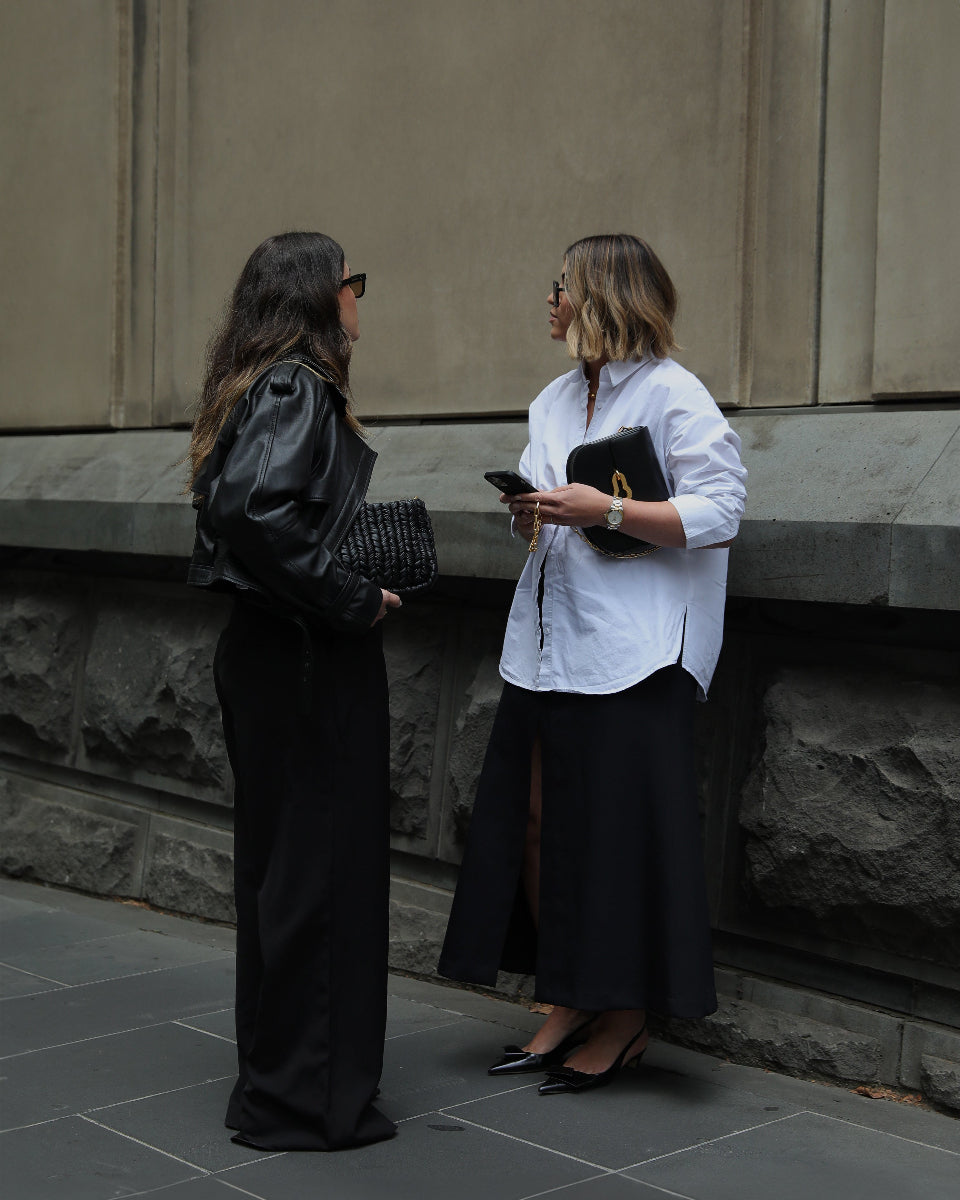 Two women standing against a stone wall, one holding a phone and the other a clutch.