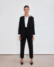 Middle aged women with brown pulled back hair wearing a  minimalistic tailored black blazer open with a crisp white shirt with the collar lifted up, neatly tucked into a pair of high-waisted black peg leg trousers which crop just above the ankle