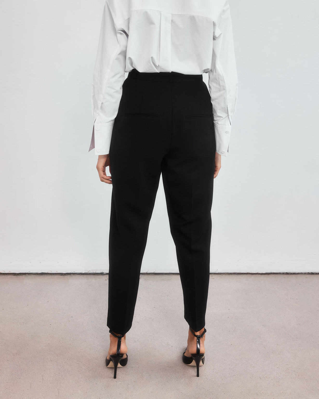 Back view of minimalistic high-waisted black peg leg trousers on a middle aged woman.
