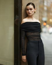 A woman wearing an off-the-shoulder, long-sleeved, semi-sheer black top with a rouched neckline.