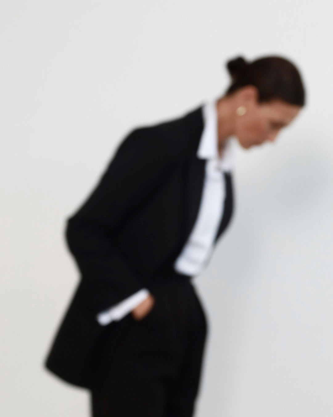 Out of focus image of a middle aged woman with pulled back dark hair wearing minimalist black suit with tailored blazer, and high waisted peg leg trousers