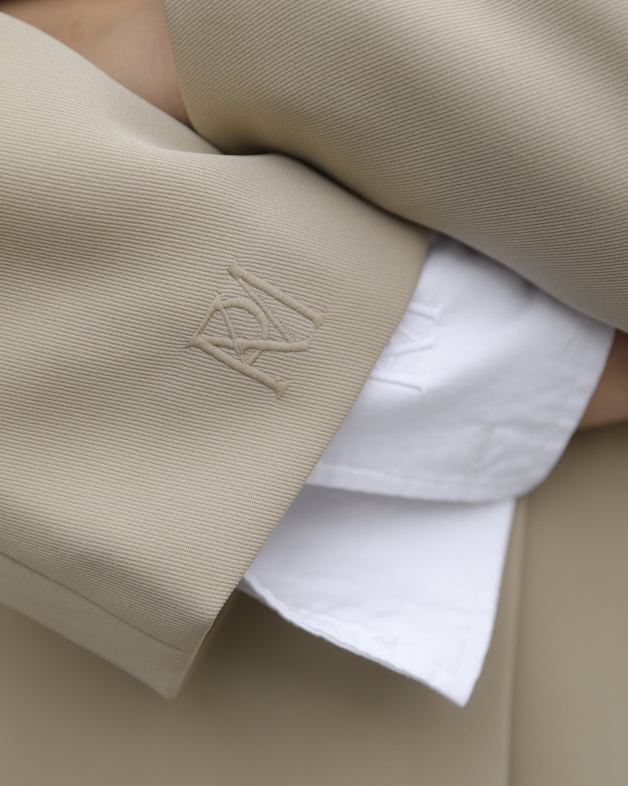 Close up image of a minimalist taupe trench coat with embroidered MRK brandmark on right sleeve, with crisp white shirt poking out from the end of the sleeve, with matching brand mark.