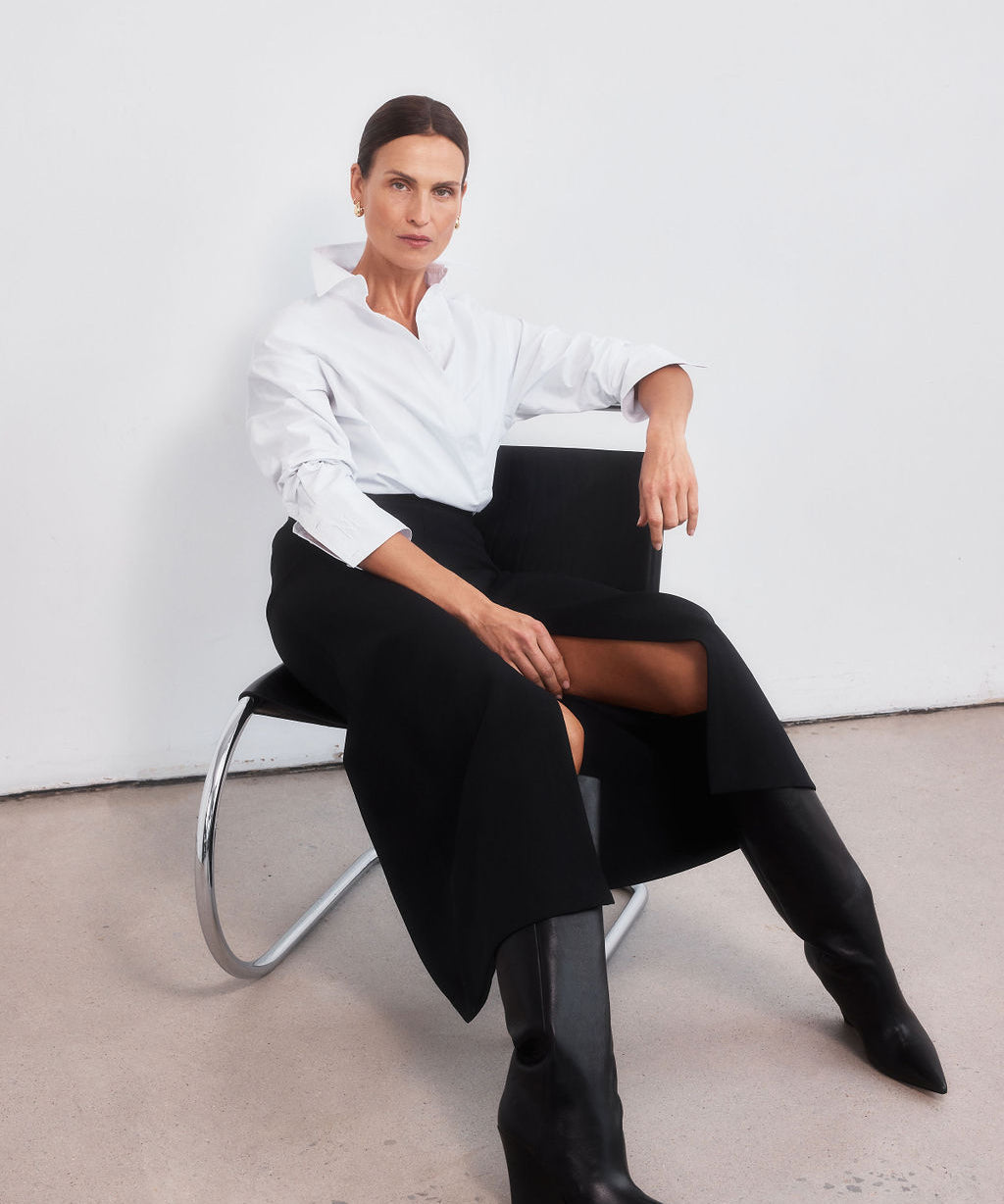 Middle aged women with brown pulled back hair standing with legs slightly apart staring directly at the camera. Styled wearing a minimalist crisp white shirt, neatly tucked into a high-waisted maxi length skirt with middle split and cream round-toe mules