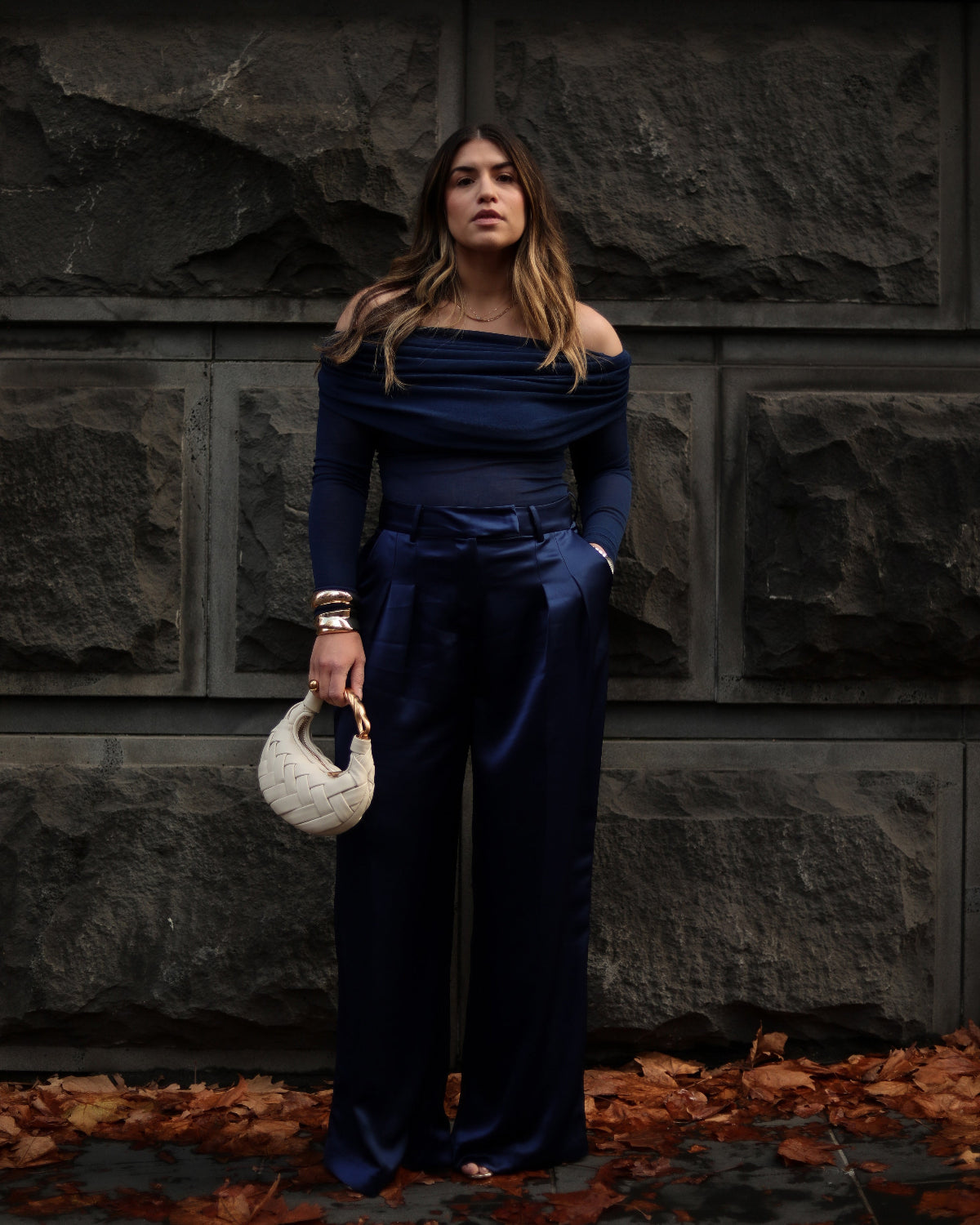 Woman in a navy blue outfit holding a textured handbag against a stone wall.