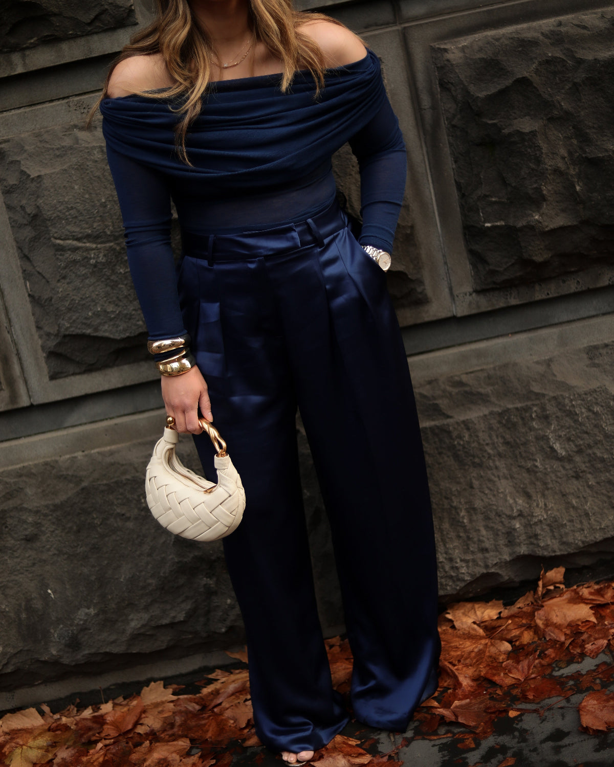 Person wearing a blue off-shoulder top and wide-leg pants, holding a white handbag against a stone wall.