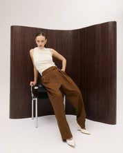 Woman in white top and brown pants posing against a wooden screen