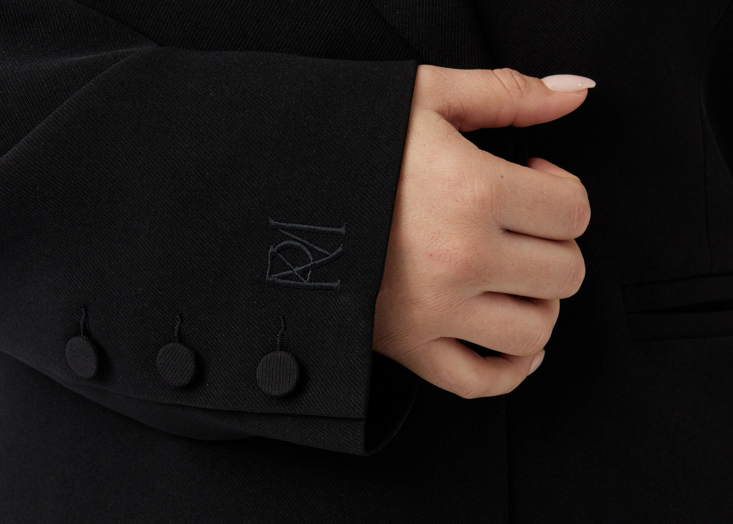 Close up image of an embroidered brand mark on the right hand sleeve of a minimalistic black blazer