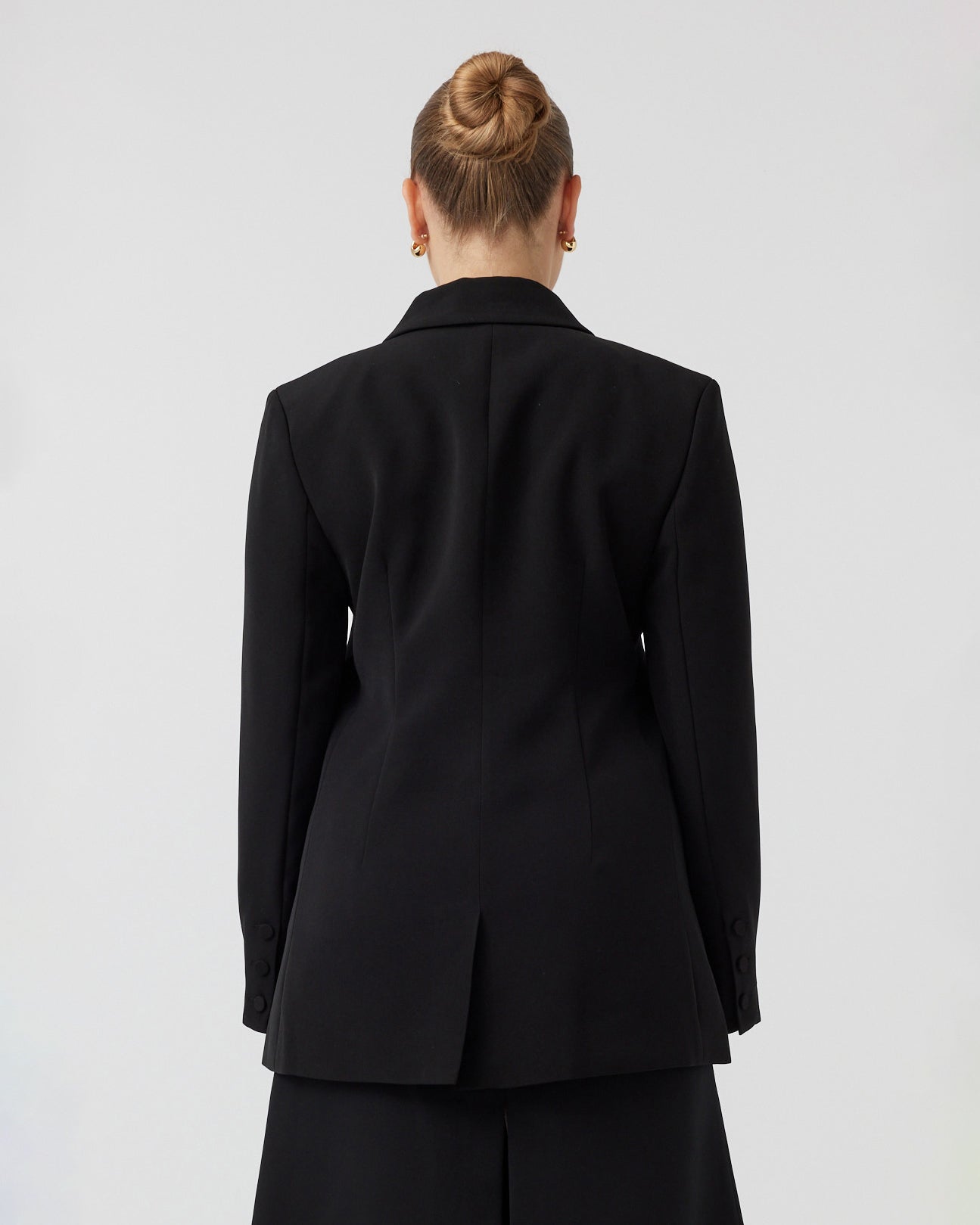 Youthful woman with pulled back dark blonde hair, with a curvaceous figure, photographed from the back wearing tailored black blazer 