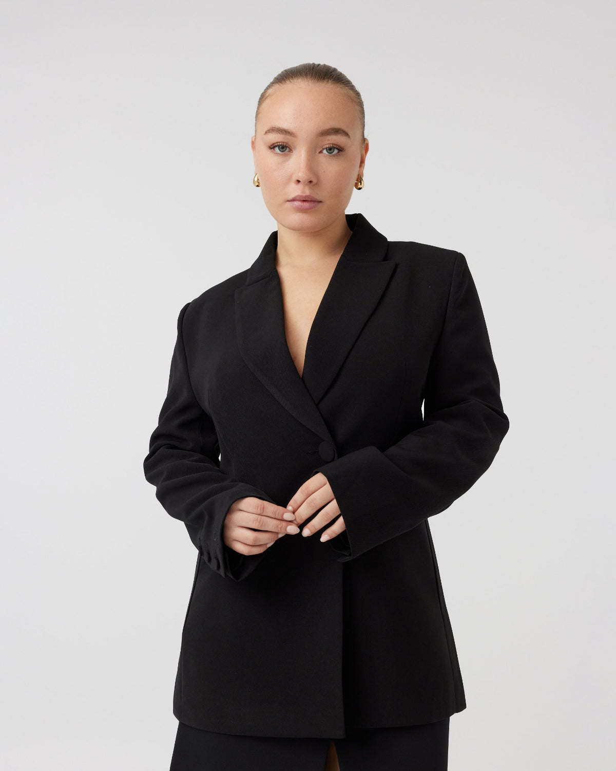 Youthful woman with pulled back dark blonde hair, with a curvaceous figure wearing tailored black blazer buttoned up, holding her hands in front of her waist