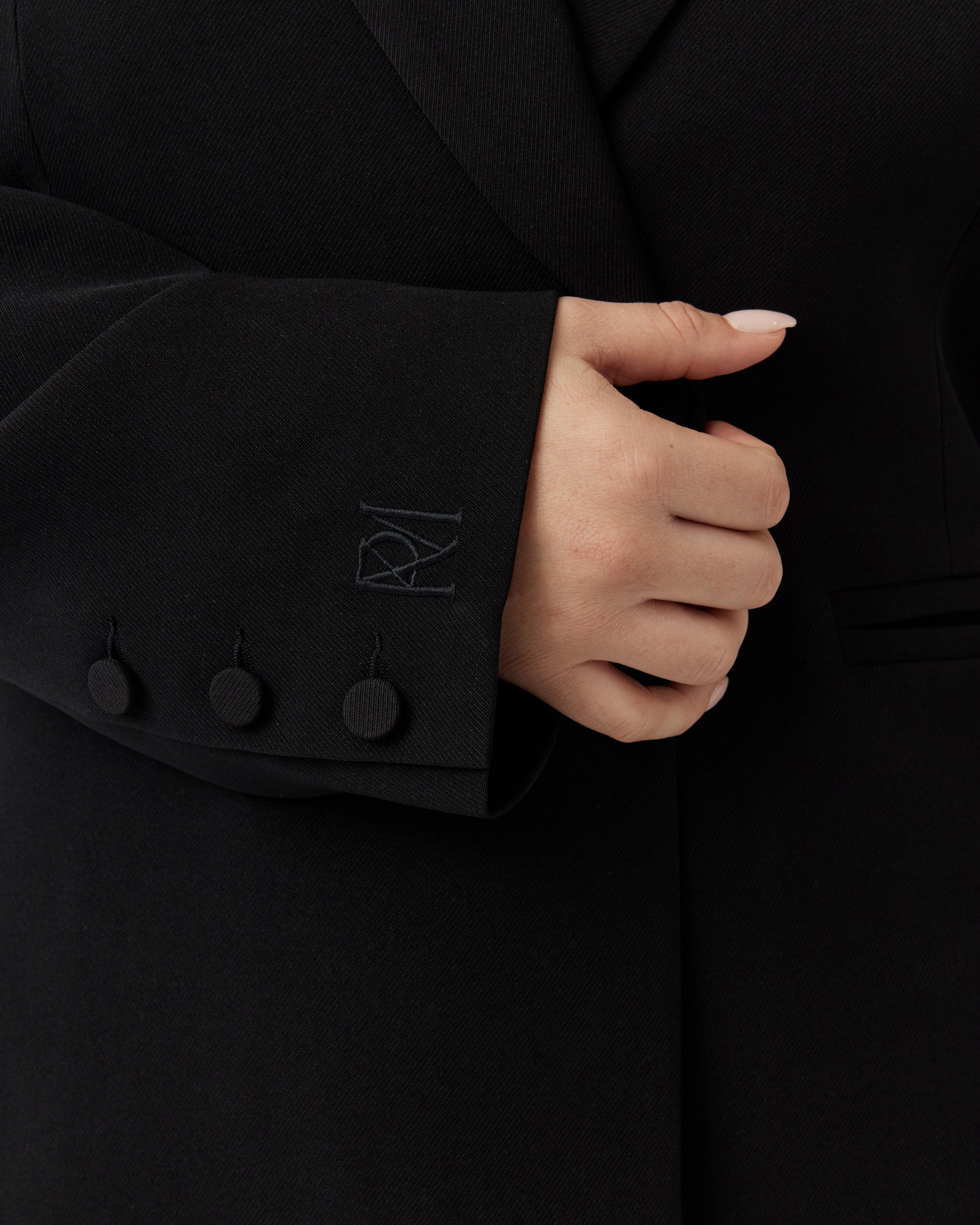 Close up image of an embroidered brand mark on the right hand sleeve of a minimalistic black blazer