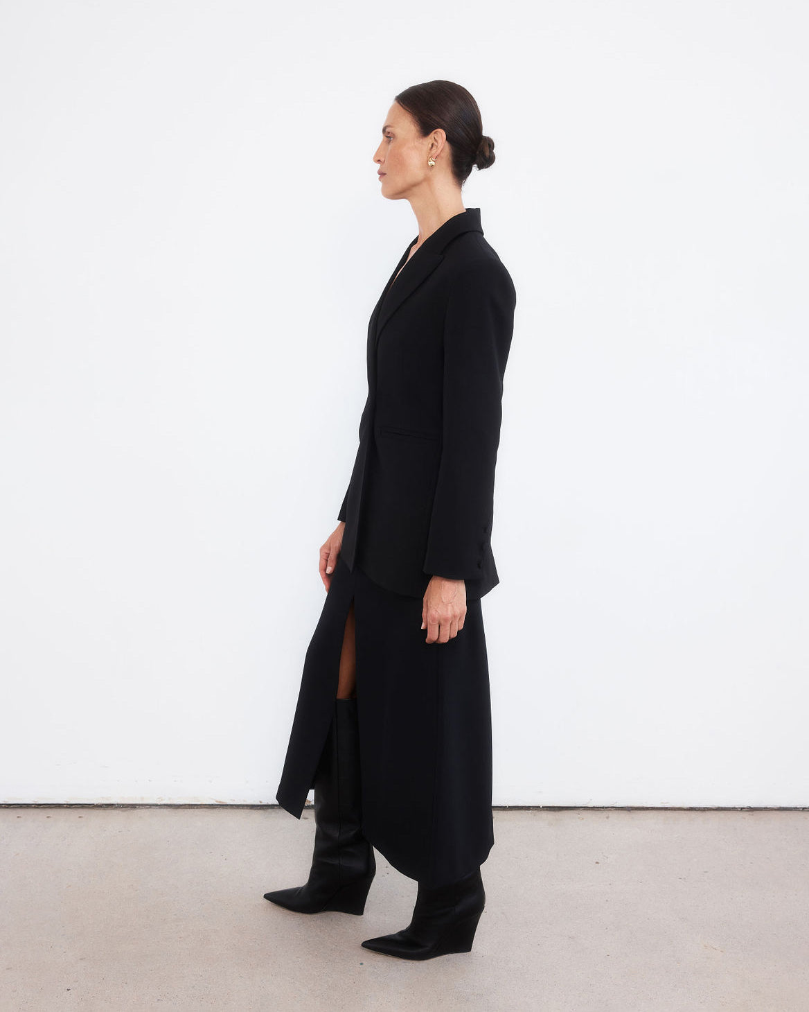 Middle aged women with brown pulled back hair, looking to the left hand side, wearing an all black look with a minimalist tailored black blazer, maxi length skirt with middle split and black wedge pointed boots