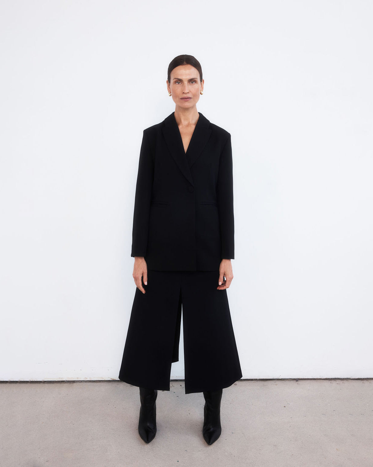 Middle aged women with brown pulled back hair wearing an all black look with a minimalist tailored black blazer, maxi length skirt with middle split and black wedge pointed boots