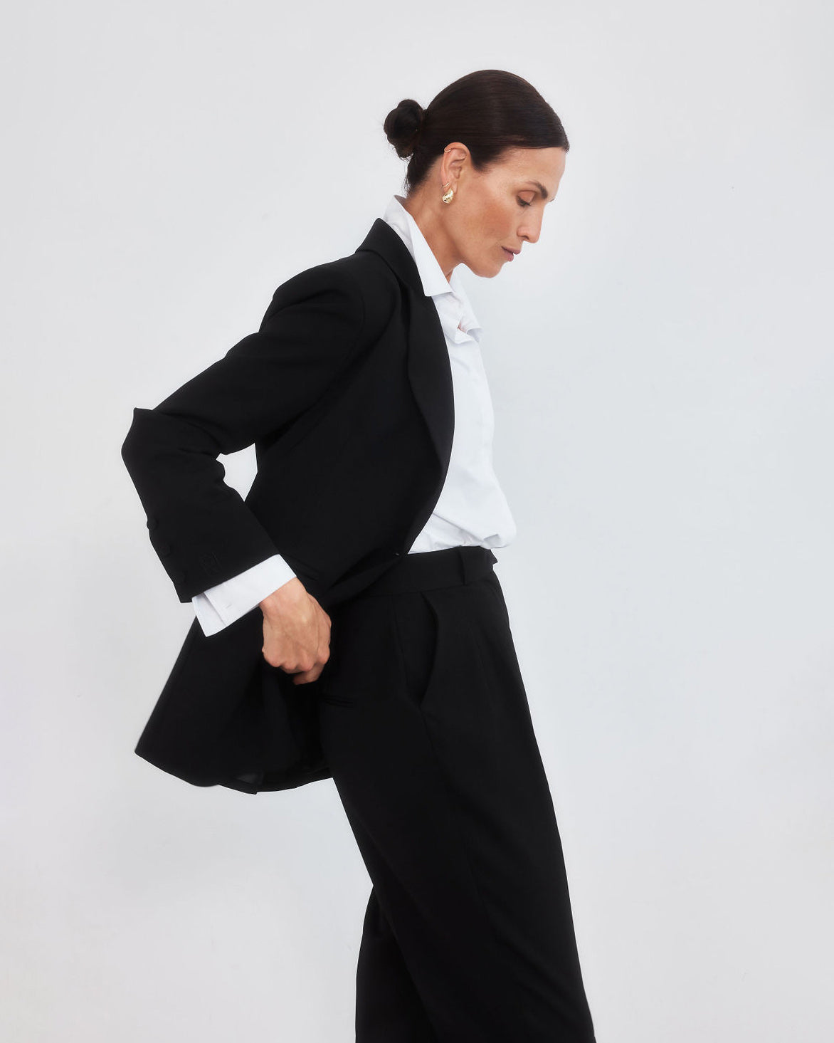 Middle aged woman with pulled back hair in a bun looking down to the right hand side, wearing a minimalist classic outfit of a tailored black blazer, crisp white shirt and high waisted black trousers