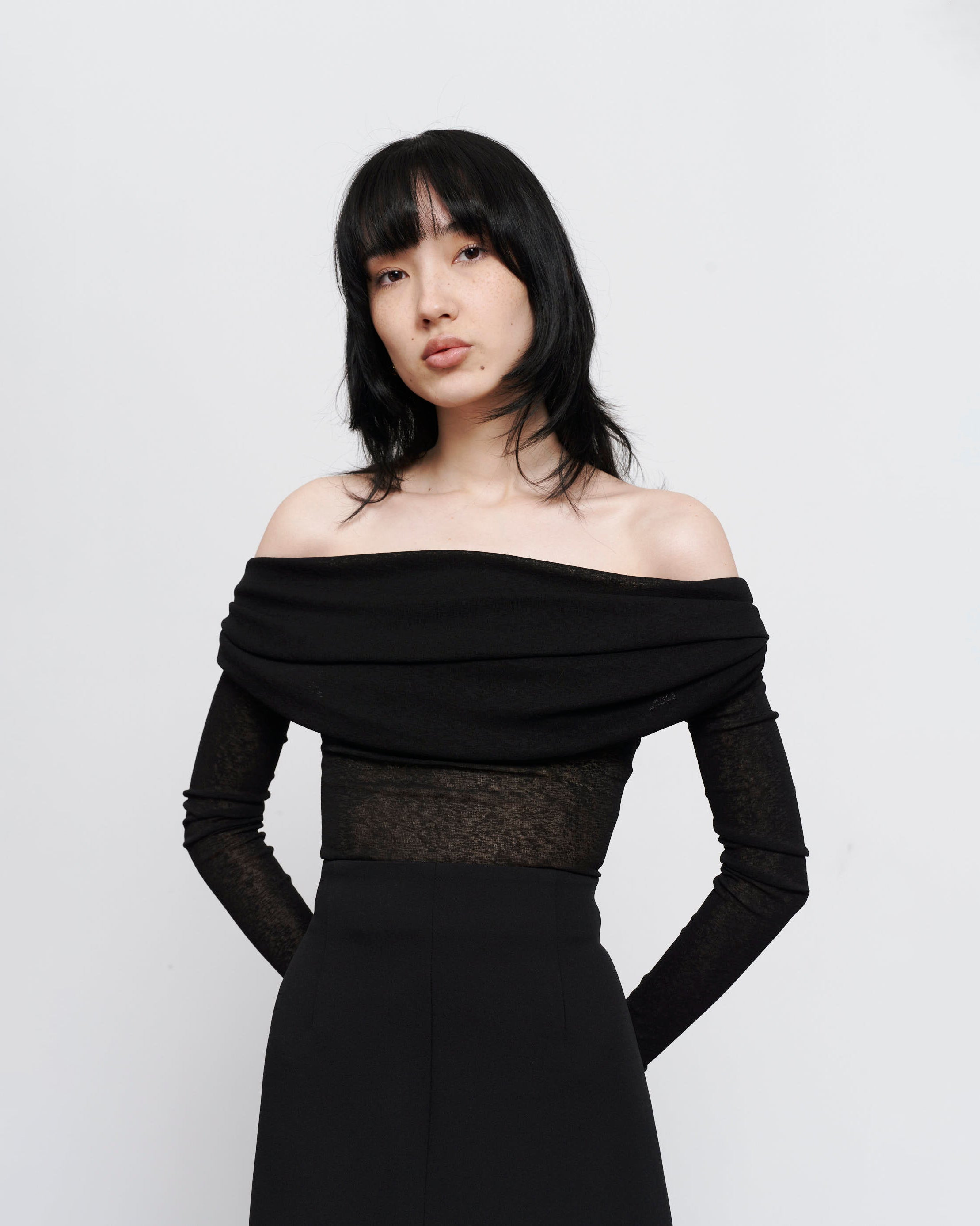 Black semi sheer, rouched off the shoulder top on women with dark hair and hands gently placed behind her back 