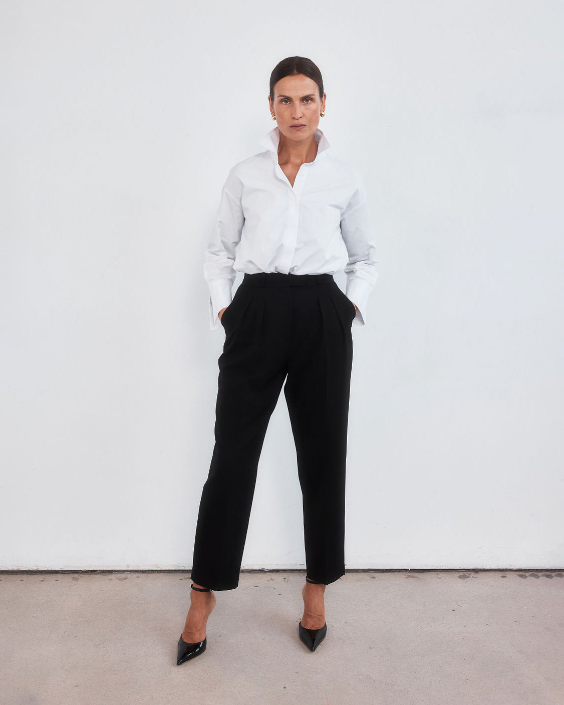Middle aged women with brown pulled back hair wearing a crisp white shirt with the collar lifted up, neatly tucked into a pair of high-waisted black peg leg trousers which crop just above the ankle