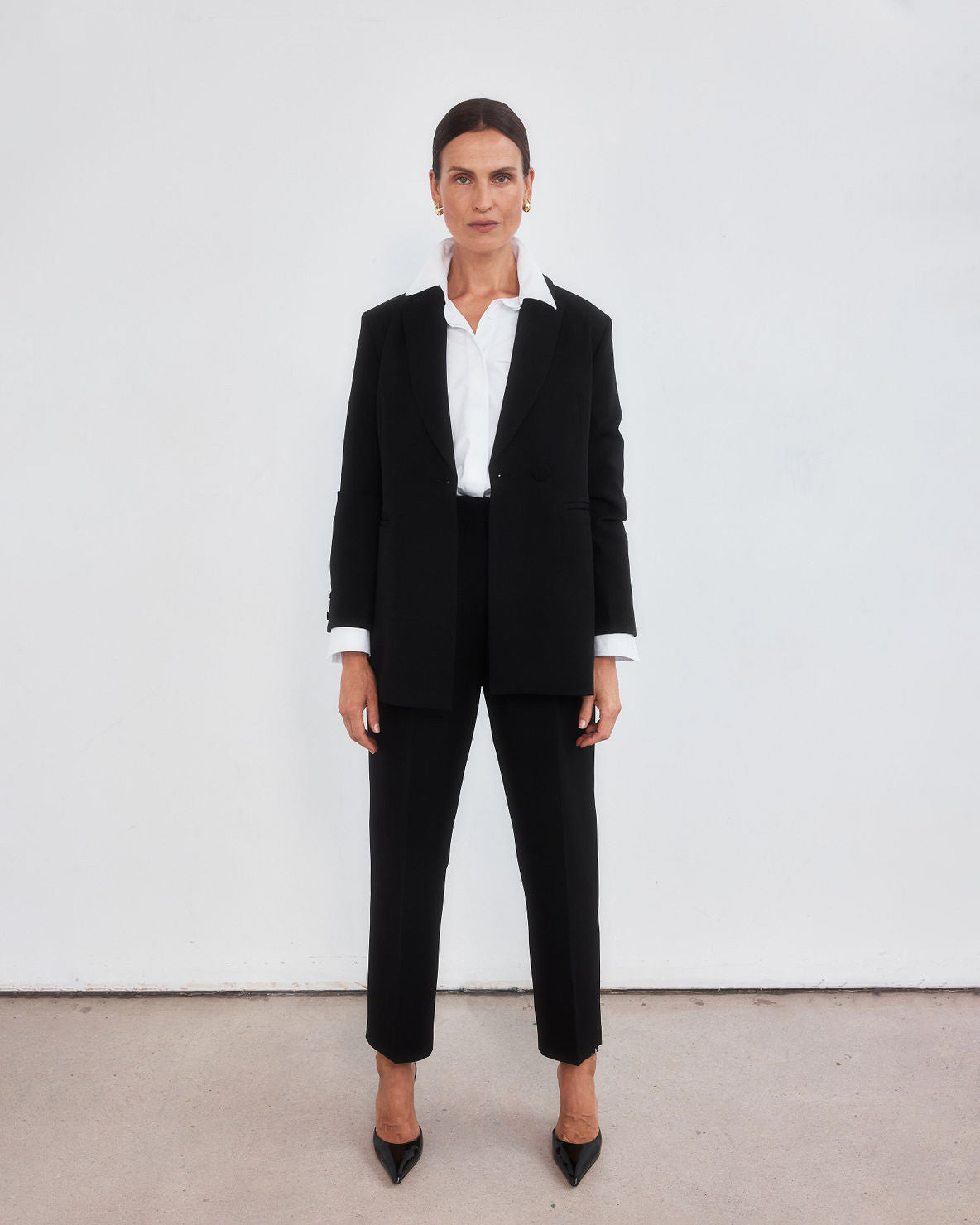Middle aged women with brown pulled back hair wearing a  minimalistic tailored black blazer open with a crisp white shirt with the collar lifted up, neatly tucked into a pair of high-waisted black peg leg trousers which crop just above the ankle