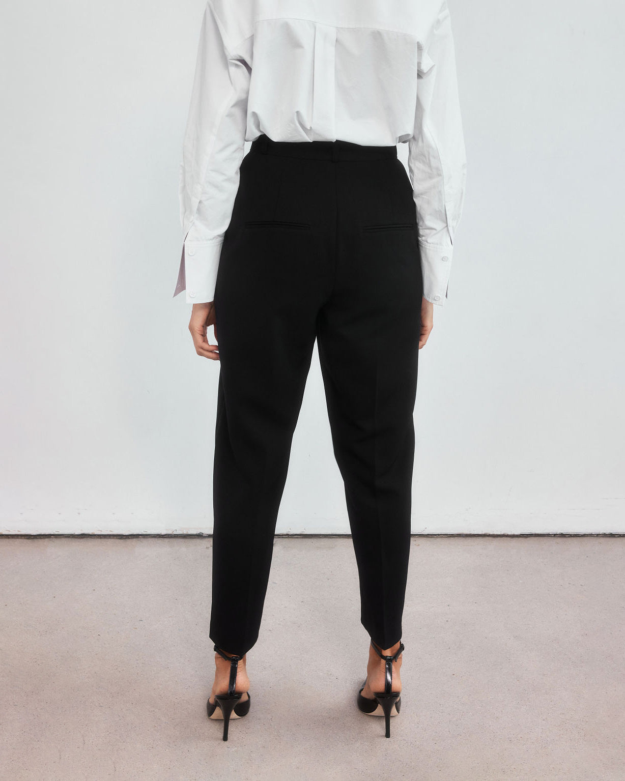 Back view of minimalistic high-waisted black peg leg trousers on a middle aged woman. 
