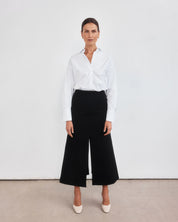 Middle aged women with brown pulled back hair standing with legs slightly apart staring directly at the camera. Styled wearing a minimalist crisp white shirt, neatly tucked into a high-waisted maxi length skirt with middle split and cream round-toe mules