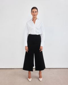 Middle aged women with brown pulled back hair standing with legs slightly apart staring directly at the camera. Styled wearing a minimalist crisp white shirt, neatly tucked into a high-waisted maxi length skirt with middle split and cream round-toe mules