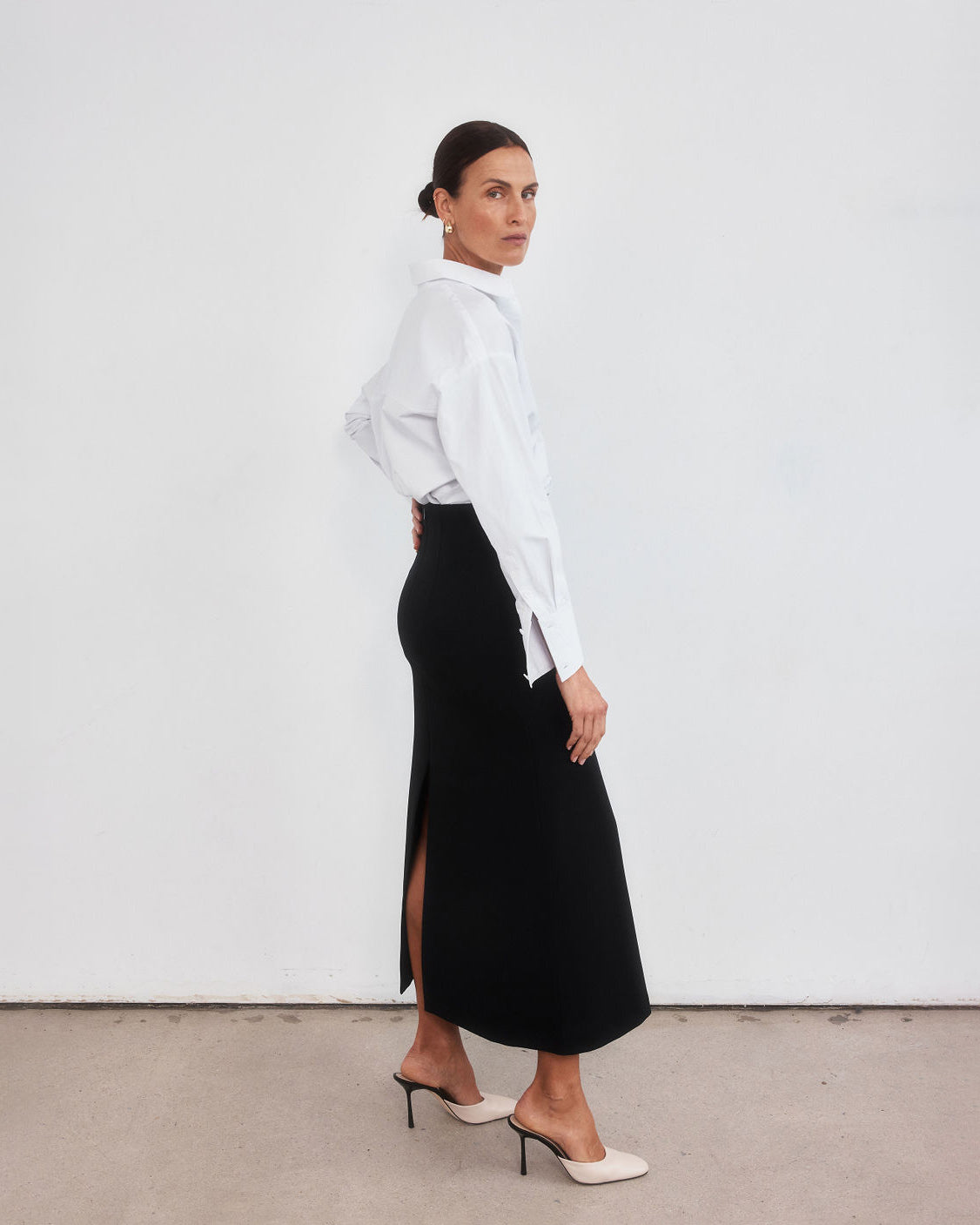 Middle aged women with brown pulled back hair standing with legs slightly apart looking over her right shoulder to the camera. Styled wearing a minimalist crisp white shirt, neatly tucked into a high-waisted maxi length skirt with middle split and cream round-toe mules