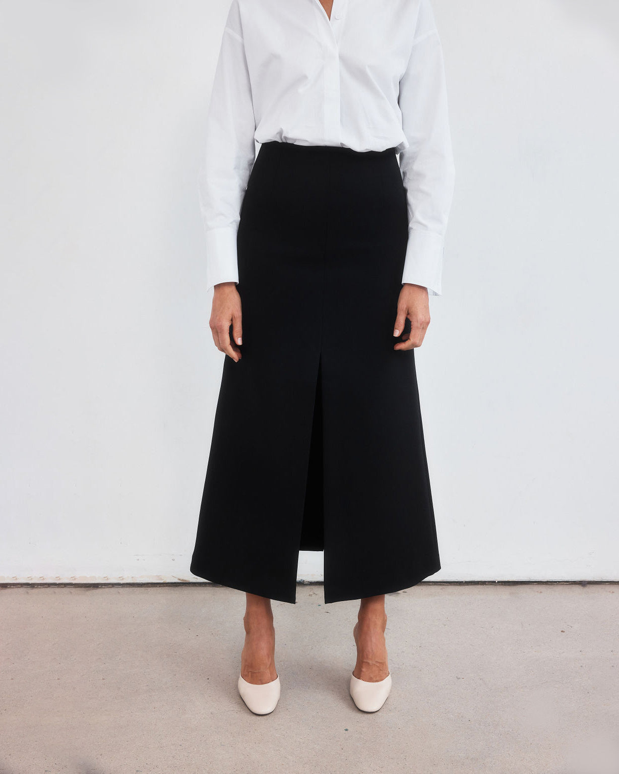 Photograph from chest height down of a woman standing with legs slightly apart staring directly at the camera. Styled wearing a minimalist crisp white shirt, neatly tucked into a high-waisted maxi length skirt with middle split and cream round-toe mules
