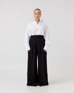 Youthful woman with dark blonde hair pulled back wearing a crisp white shirt neatly tucked into a pair of minimalistic black high waisted wide leg trousers.