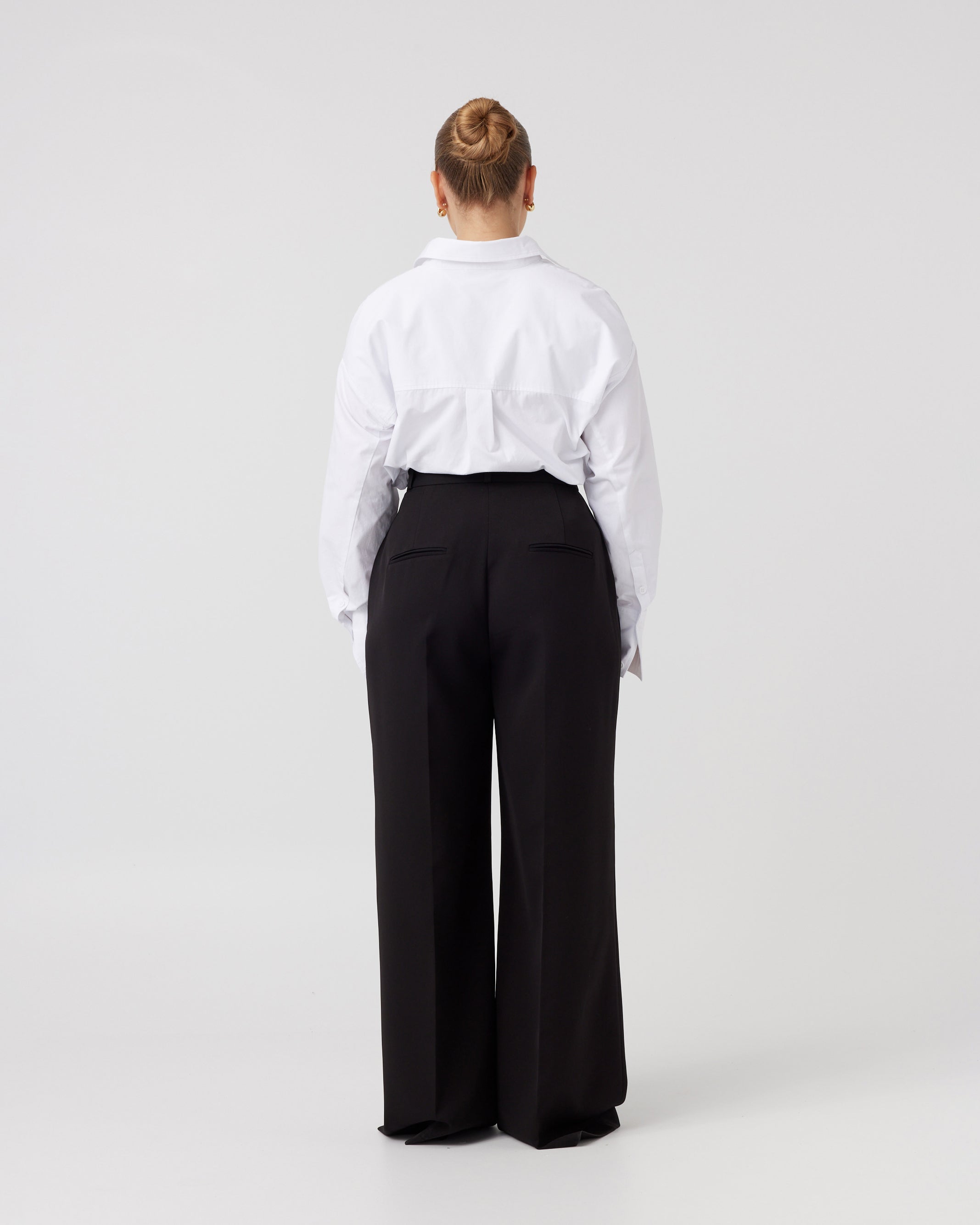 Photograph of the back of a youthful woman with dark blonde hair pulled back wearing a crisp white shirt neatly tucked into a pair of minimalistic black high waisted wide leg trousers.