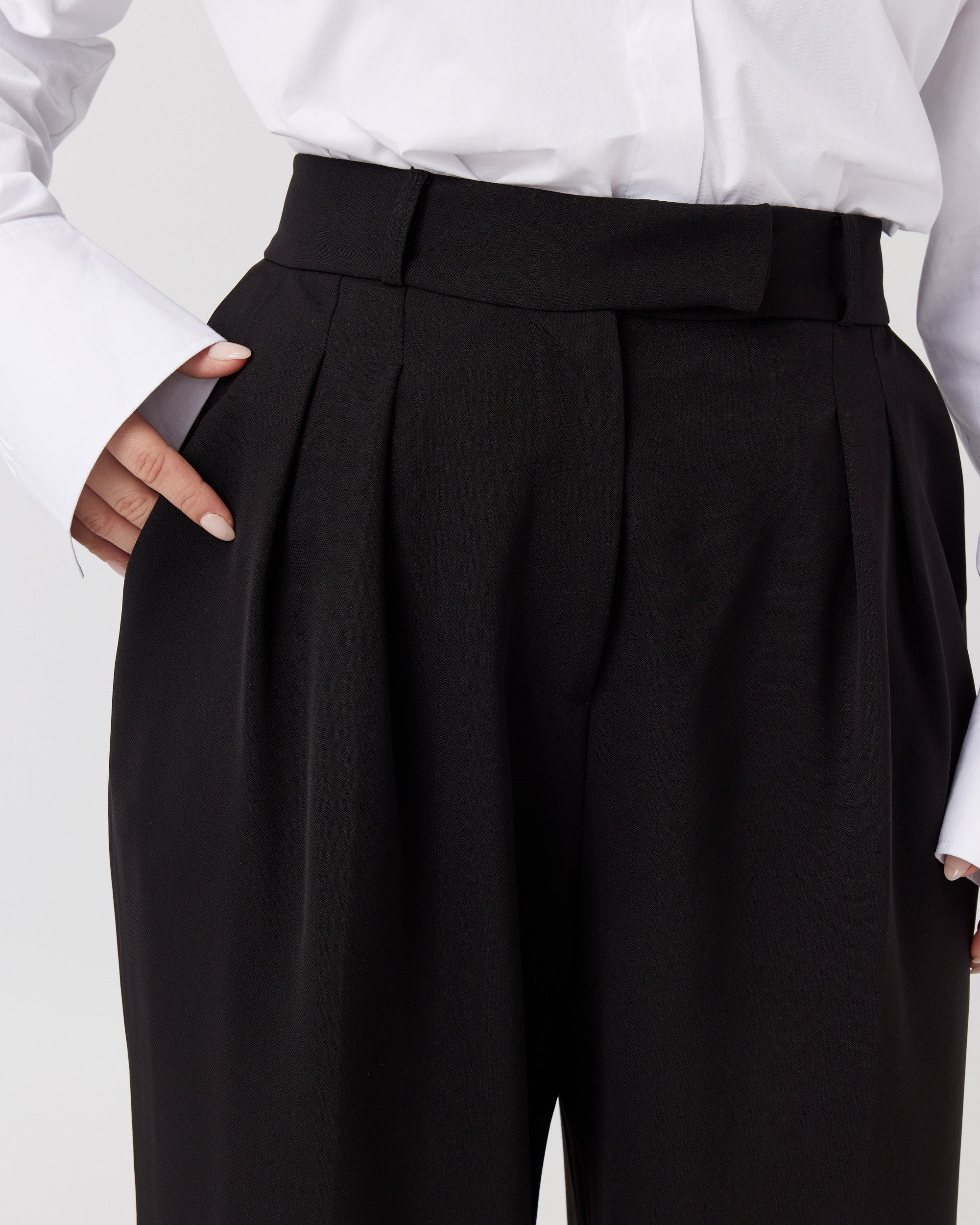 Close up image of the waist band of a curvaceous women wearing a pair of minimalistic high waisted wide leg trousers in black. Waistband shows darting and pleating on either side which creates a elegant drape of the fabric over the body