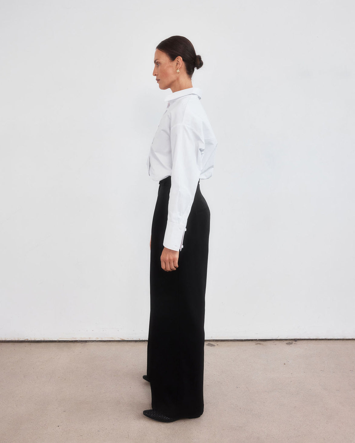 Middle aged woman with pulled back dark brown hair wearing a crisp white shirt neatly tucked into a pair of minimalistic black high waisted wide leg trousers. 