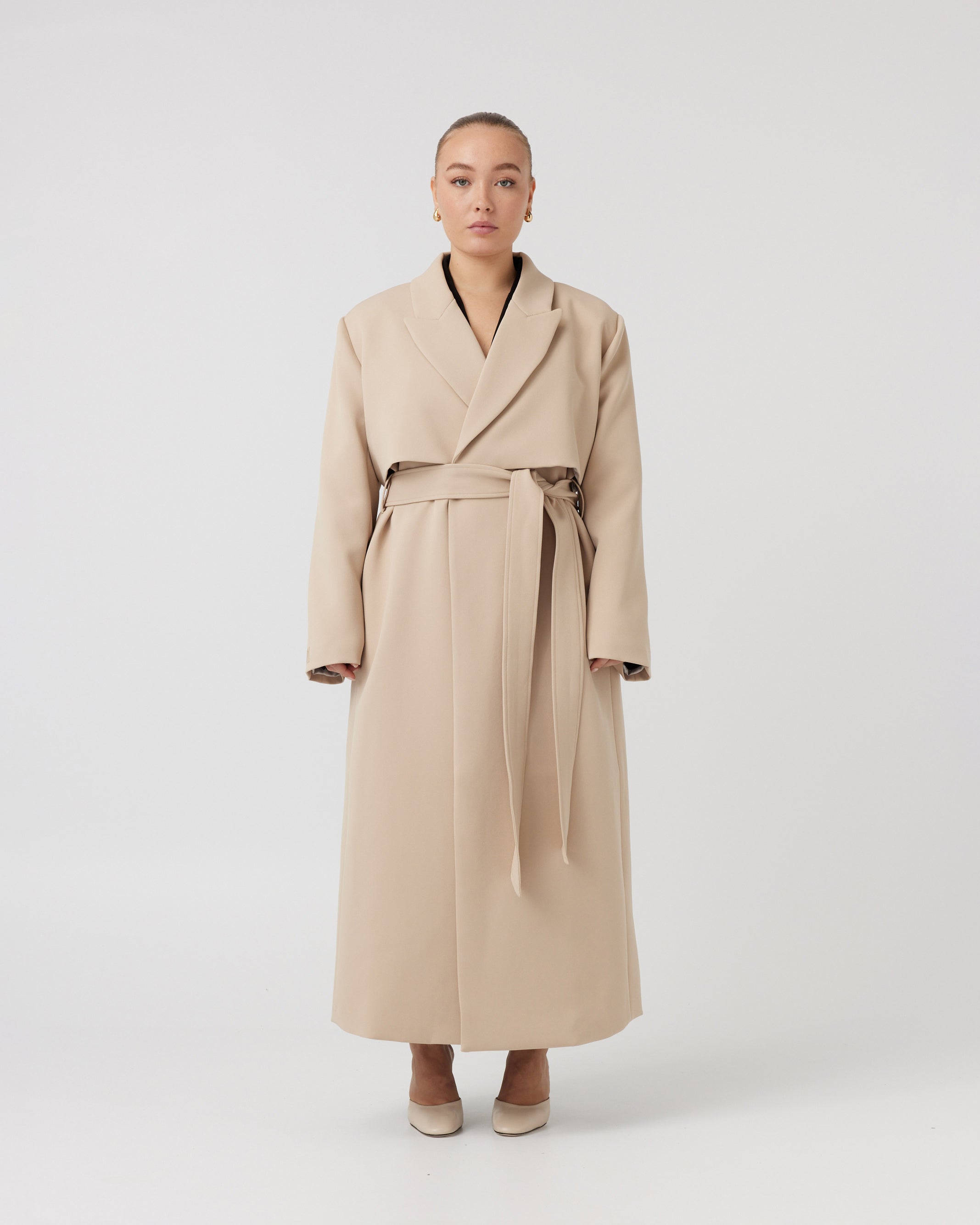 Wide angle image of a curvaceous woman with dark blonde hair wearing a minimalistic trench coat in taupe tied up around her waist with a matching taupe waist tie. Trench coat flows over her body and cuts just above the ankle.