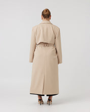 Wide angle back image of a curvaceous woman with dark blonde hair wearing a minimalistic trench coat in taupe tied up around her waist with a matching taupe waist tie. Trench coat flows over her body and cuts just above the ankle.