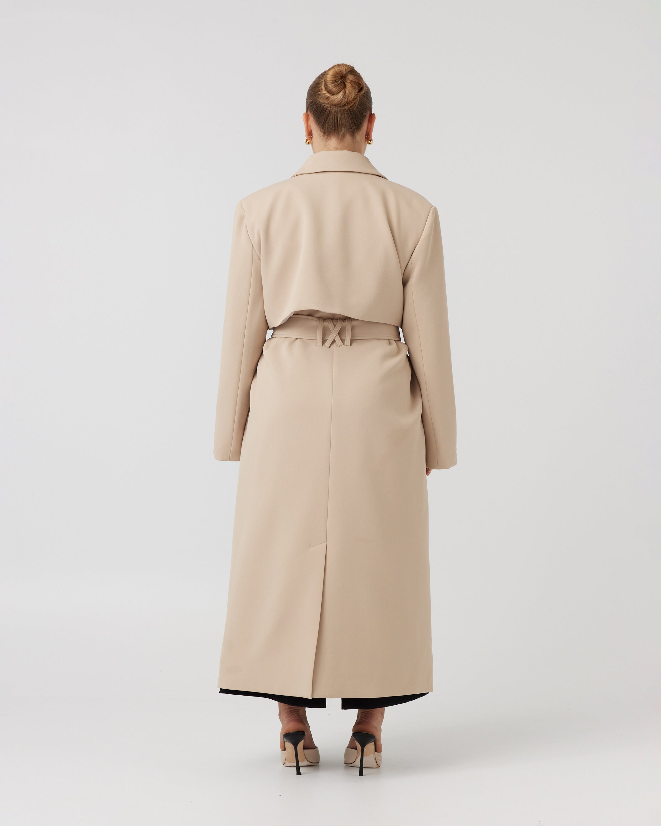 Wide angle back image of a curvaceous woman with dark blonde hair wearing a minimalistic trench coat in taupe tied up around her waist with a matching taupe waist tie. Trench coat flows over her body and cuts just above the ankle.