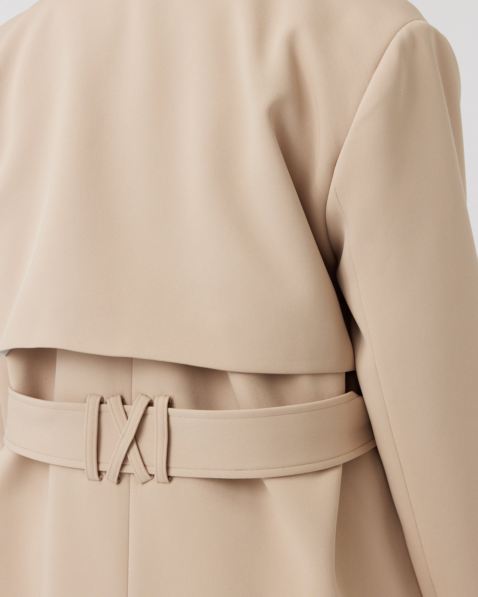 Close up detail of a curvaceous woman wearing a minimalistic trench coat in taupe with a waist tie detail at the back of the coat. 
