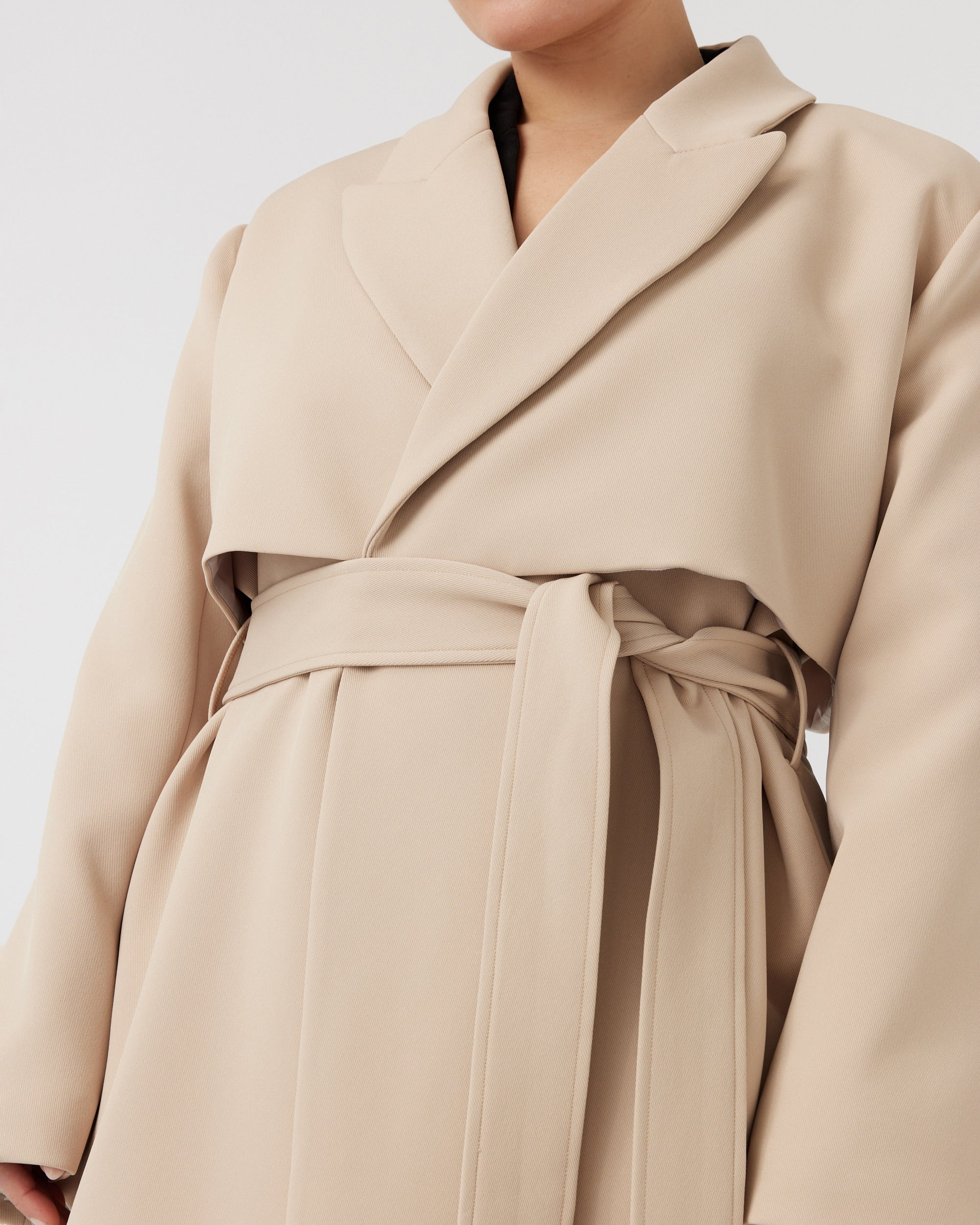 Detail image of a curvaceous woman wearing a minimalistic trench coat in taupe with storm flap detail, tied up around her waist with a taupe waist belt