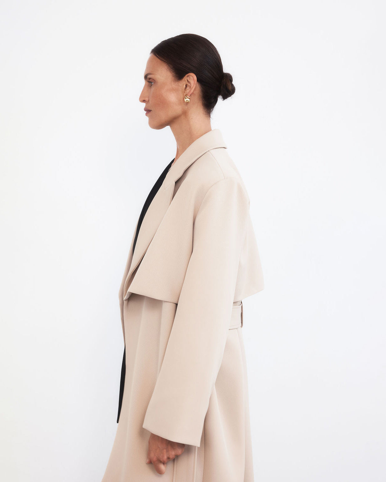 Mid photograph crop of a middle aged women with pulled back dark hair facing the left wearing a minimalistic trench coat in taupe with strong lapels and storm flap detail