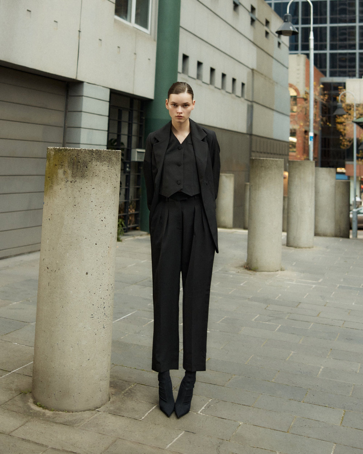 A person standing on a sidewalk wearing black tailored trousers and a black top, with concrete columns and a building in the background.