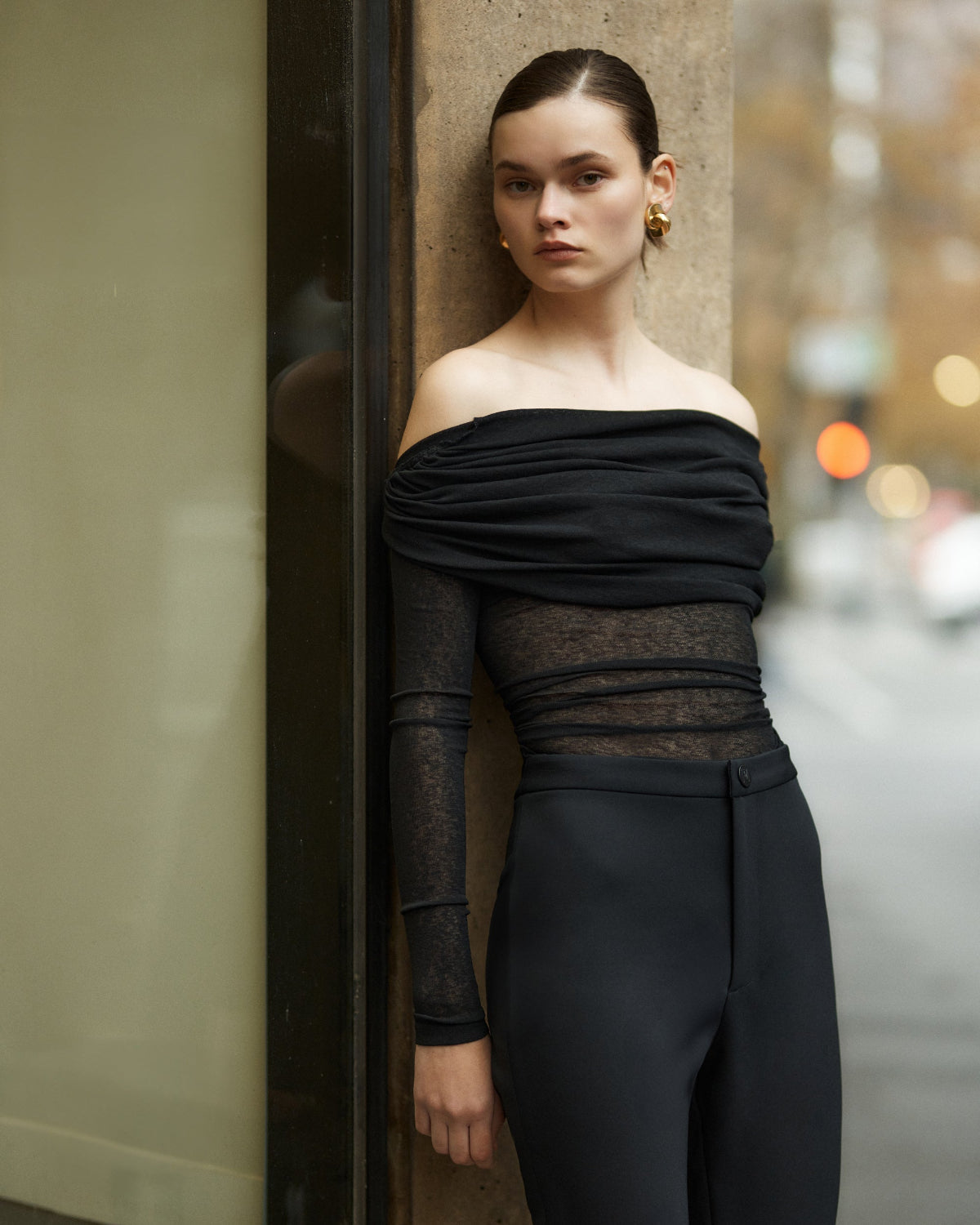 A woman wearing an off-the-shoulder, long-sleeved, semi-sheer black top with a rouched neckline.
