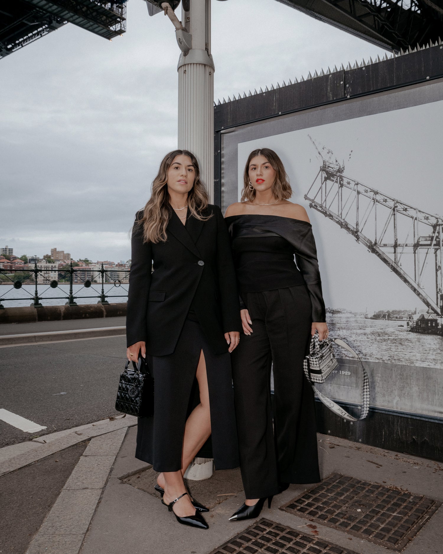 Identical twin sisters standing underneath the Sydney Harbour Bridge both wearing all black minimalist outfits staring directly at the camera. Both holding Lady Dior handbags
